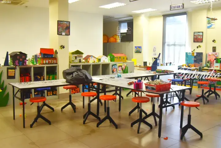 Bright classroom with tables and orange stools, featuring children’s artwork and cardboard houses on shelves.