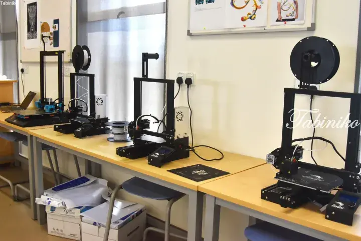Multiple 3D printers on a wooden table in a workspace with filament spools and printed designs on the wall.