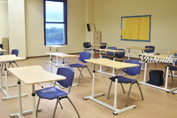 Bright classroom with adjustable height desks and blue chairs arranged in rows near a large window.