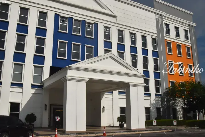 Modern hotel entrance with white portico, blue and orange facade panels, and a valet standing by the driveway.