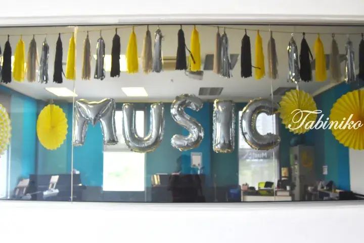 Silver foil balloons spelling MUSIC hang in a bright music studio with yellow paper fans and tassels.