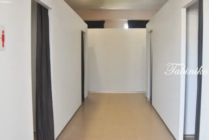 Clean hallway with white walls and beige tiled flooring, featuring dark curtains and a red male restroom sign.