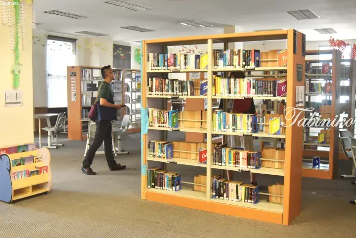 A man walks through a bright library with wooden bookshelves organized by alphabet and a children’s reading corner.