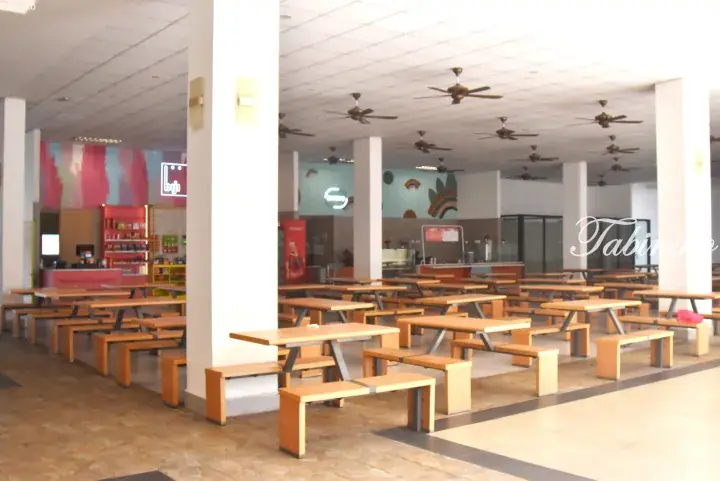 Spacious cafeteria with wooden tables and benches, ceiling fans, and colorful wall murals.