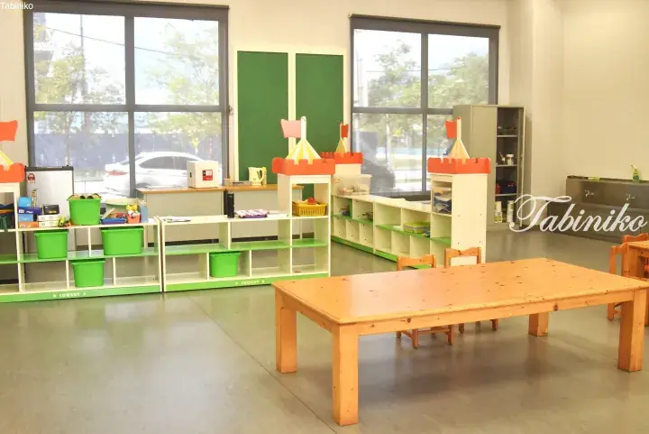 Bright kindergarten classroom with wooden table, green storage shelves, and castle-themed dividers near large windows.