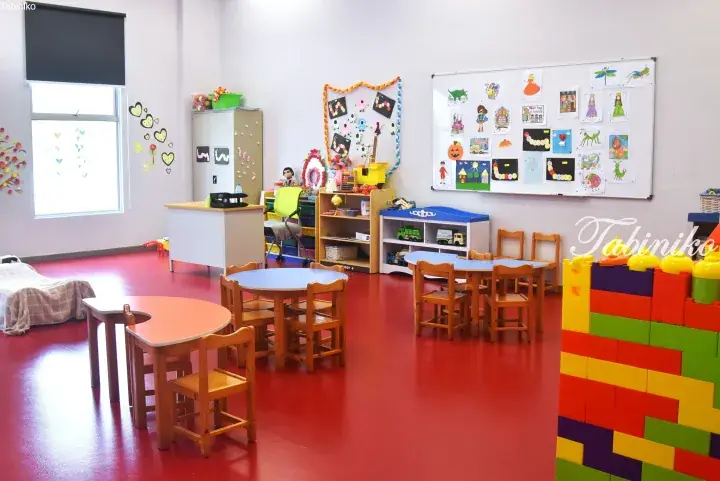 Bright kindergarten classroom with red flooring, small wooden tables, colorful wall displays, and a large Lego block structure.