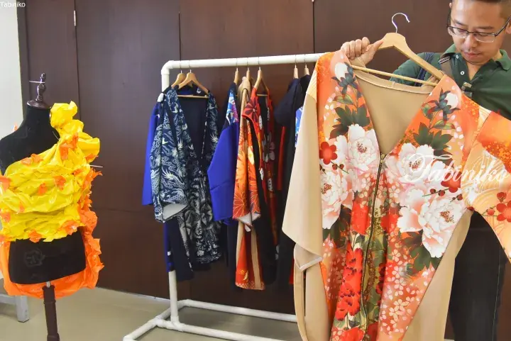 Man holding a floral kimono jacket while a rack displays various Japanese garments and a mannequin wears a yellow dress.