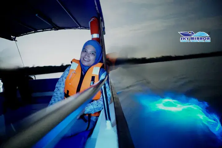 Smiling woman in life jacket on boat at night watching glowing blue bioluminescent water in Malaysia
