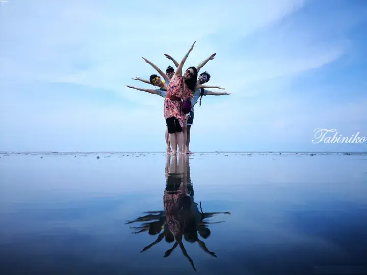 Group of friends posing with arms outstretched on a reflective tidal flat, creating a mirror effect under a blue sky.