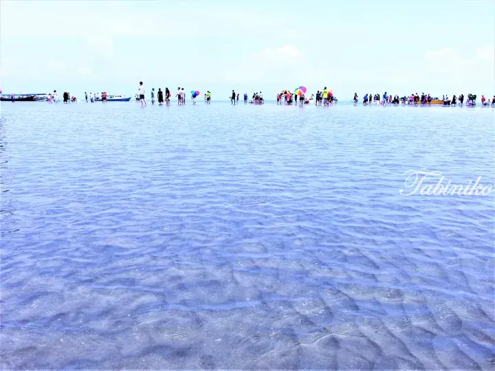 Tourists wading in shallow clear water at a Malaysian beach with boats and colorful umbrellas in the distance.