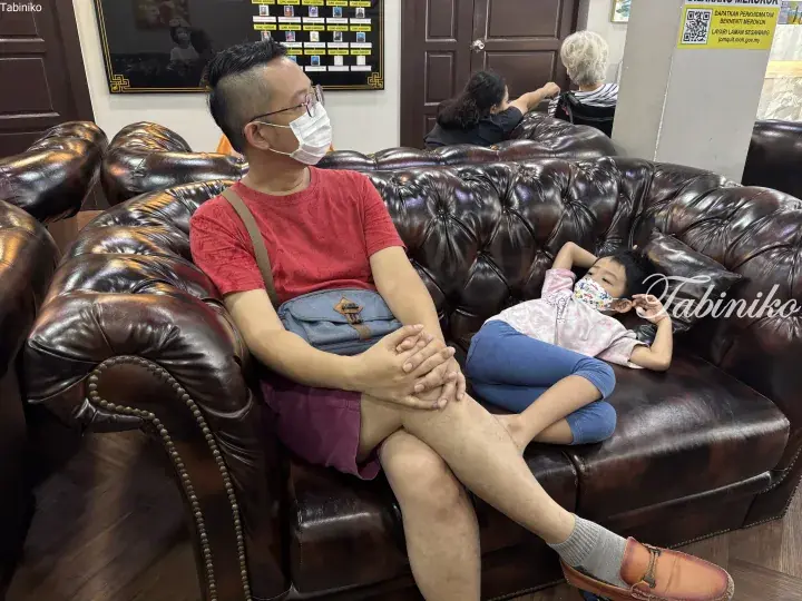 A man and child relax on a brown leather Chesterfield sofa in a clinic waiting area.