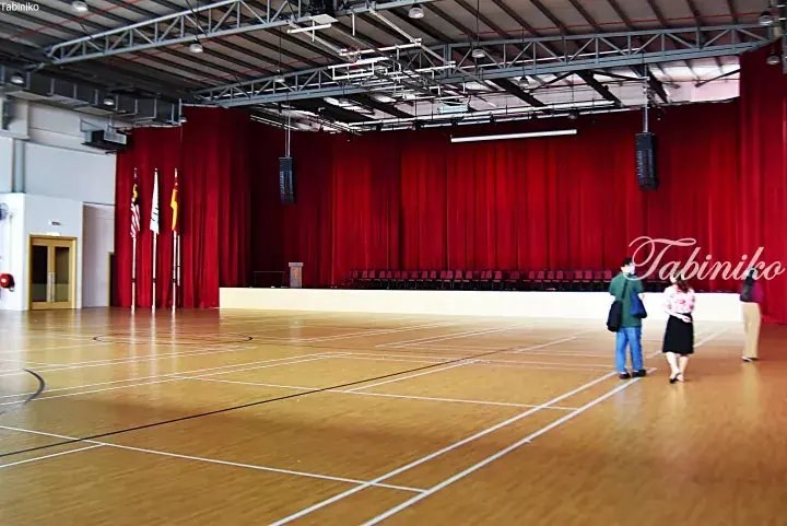 Spacious indoor hall with wooden floor, red curtains, and Malaysian flags, likely a school or community center in Malaysia.