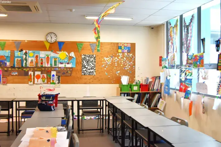 Bright classroom with student artwork on cork boards and windows, featuring desks, chairs, and a clock.