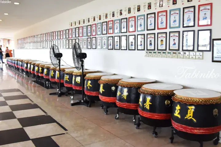 Row of large traditional drums labeled with Chinese characters displayed in a school hallway with framed certificates on the wall.