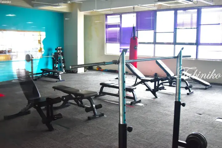 Gym area with weight benches, a barbell rack, and a red punching bag near large windows with purple blinds.
