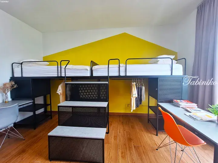 Modern hostel room with yellow accent wall, black metal bunk beds, and study desks.