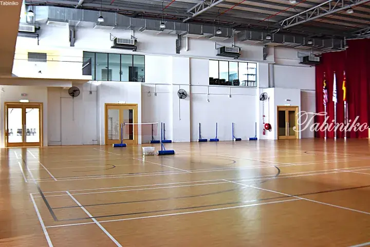 Spacious indoor sports hall with wooden flooring, badminton nets, and a stage with Malaysian flags.