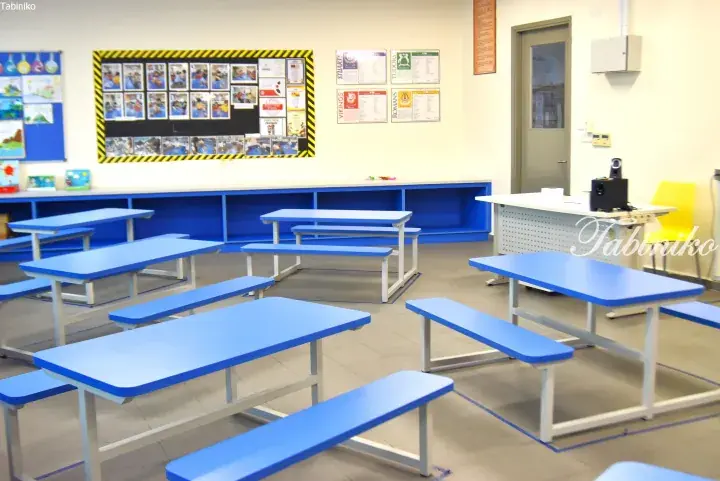 Bright classroom with blue tables and benches, display boards, and educational posters.