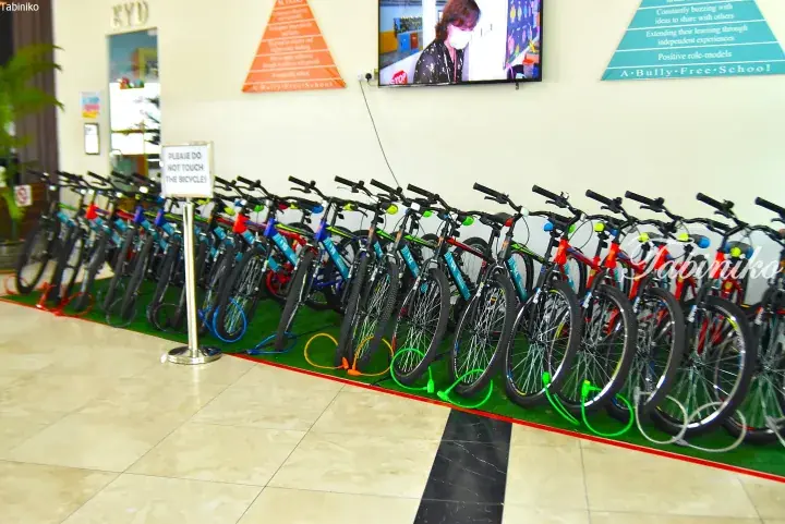 Colorful bicycles lined up on artificial grass inside a school lobby with a ‘Please Do Not Touch’ sign.