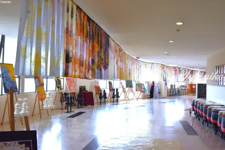 Colorful fabric art installation and paintings displayed in a bright gallery hall with giant chess pieces and drums.