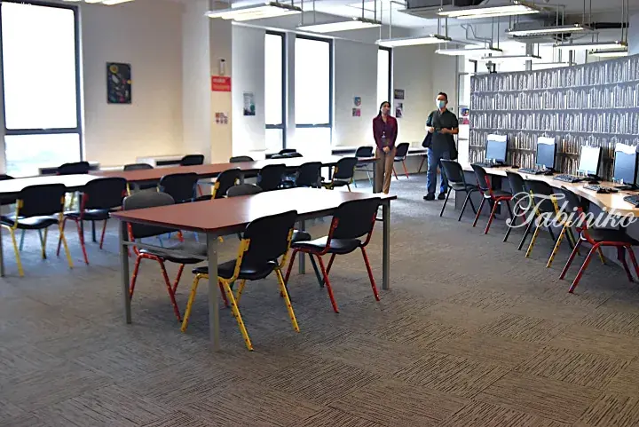 Modern library interior with study tables, colorful chairs, and computer stations near a bookshelf wall.