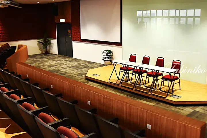 Modern lecture hall with tiered seating, a wooden stage, and a large projection screen for presentations.