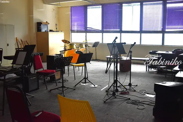 A spacious music practice room with drum kits, amplifiers, microphones, and colorful chairs near large windows.