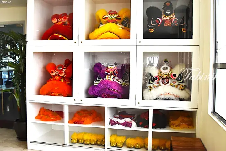 Colorful lion dance heads displayed in white glass cabinets at a Malaysian venue.