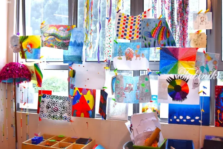 Colorful children’s artwork and crafts hanging from strings in a bright classroom window display.