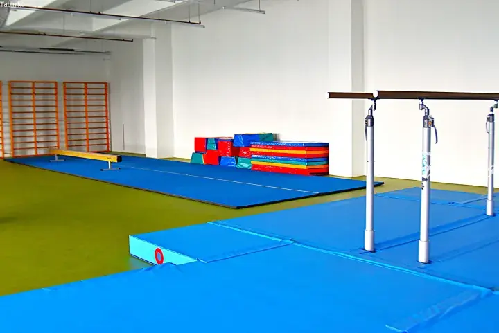 Bright indoor gymnastics facility with blue mats, parallel bars, balance beam, and colorful foam blocks.