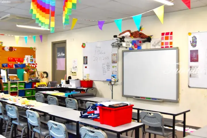Bright classroom with student desks, whiteboards, and colorful decorations hanging from the ceiling.