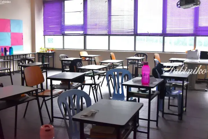 Empty classroom with purple blinds, wooden desks, and blue plastic chairs in a Malaysian school setting.