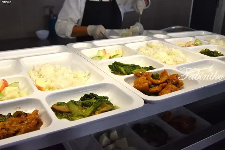 Rows of compartmentalized meal trays filled with rice, vegetables, and meat being prepared by a worker in a kitchen.