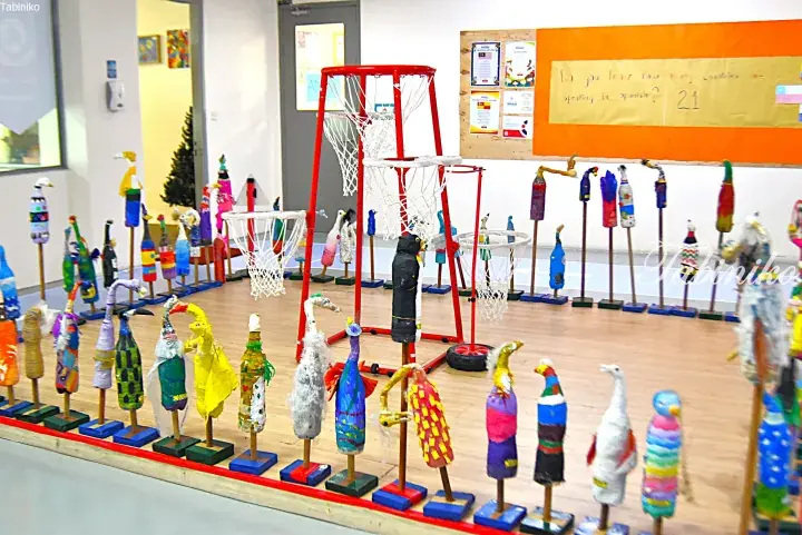 Colorful recycled bird sculptures displayed in a school hall with basketball hoops and a Spanish language bulletin board.