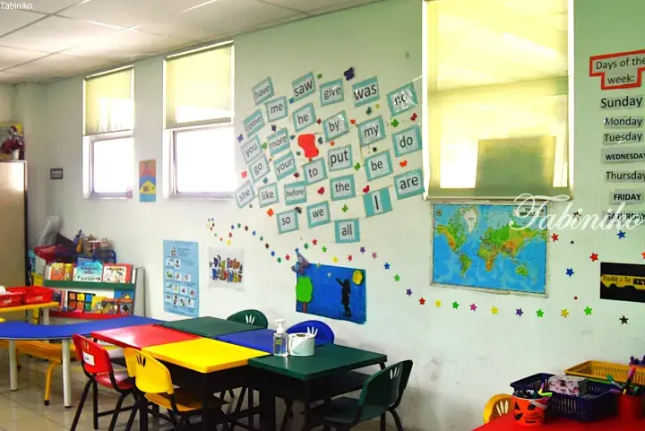 Colorful classroom with child-sized tables, chairs, and educational wall displays including a world map and English vocabulary cards.