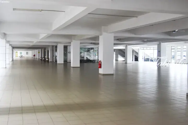 Spacious, empty hall with white pillars and tiled flooring, featuring a red fire extinguisher and ceiling fans.