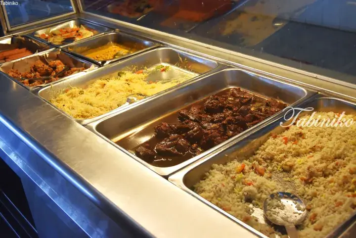 Buffet display with stainless steel trays of rice, stewed meat, noodles, and fried chicken.