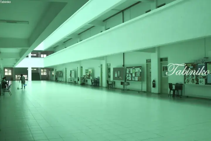 Spacious school corridor with white tiled floors, bulletin boards, and a student sweeping near the entrance.