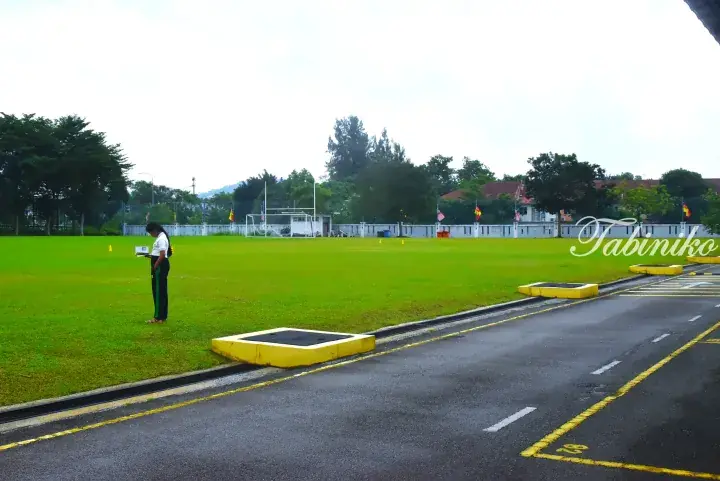 Student in uniform reads on a school sports field with goalposts and flags visible in the background.
