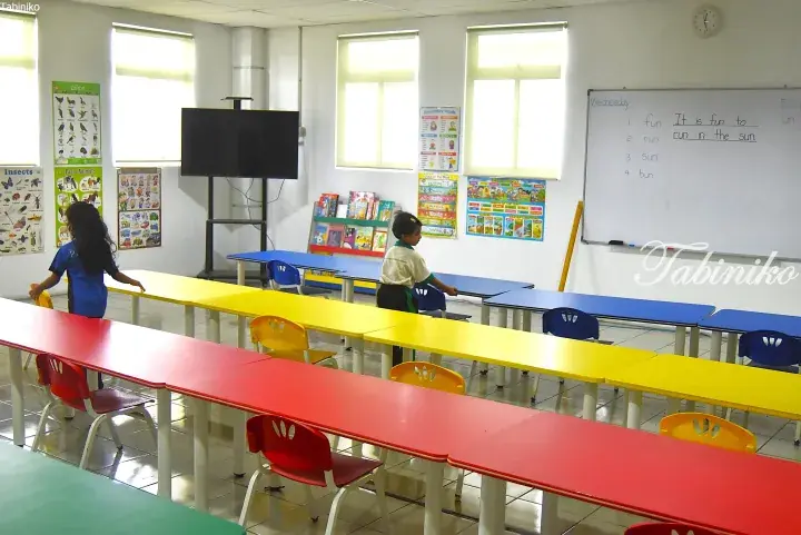 Students in a bright classroom with colorful tables, educational posters, and a whiteboard.