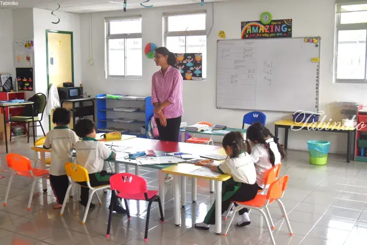 Teacher instructs young students in a bright classroom with colorful furniture and a whiteboard at an international school in Malaysia.