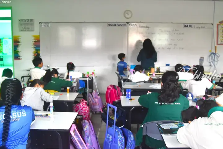 Students in green and blue uniforms sit at desks in a classroom while a teacher writes on a whiteboard.
