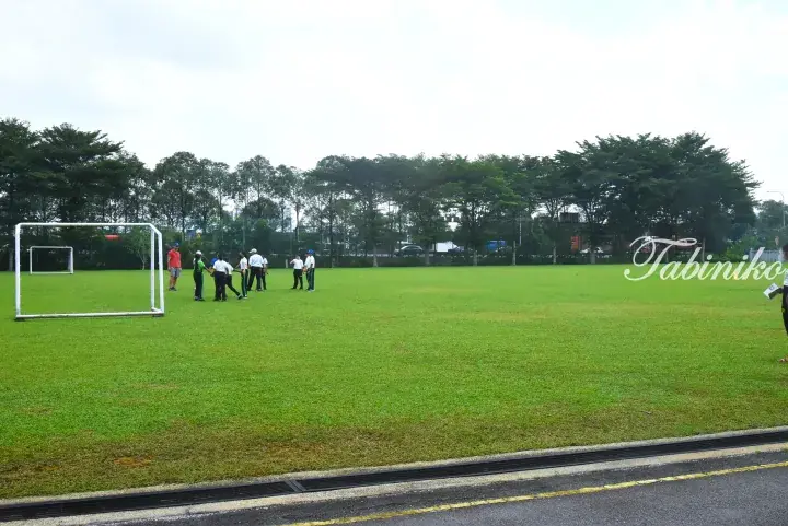 Students in uniforms playing soccer on a green field with trees in the background at a Malaysia school or park.