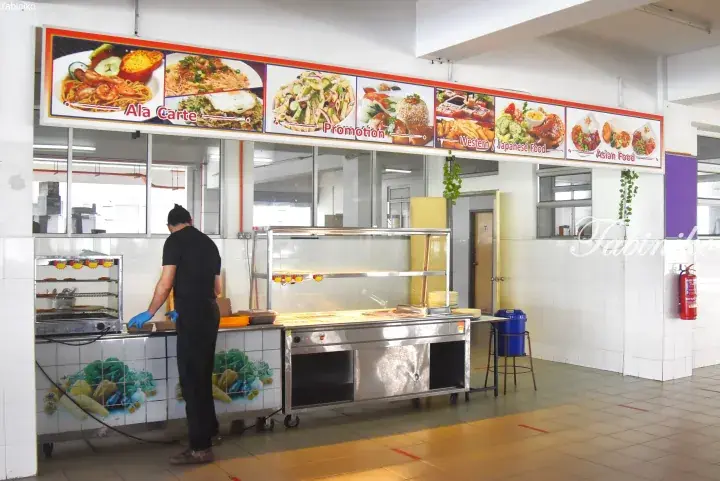 Staff preparing food at a cafeteria counter with a menu board displaying Ala Carte, Promotion, Western, and Asian options.