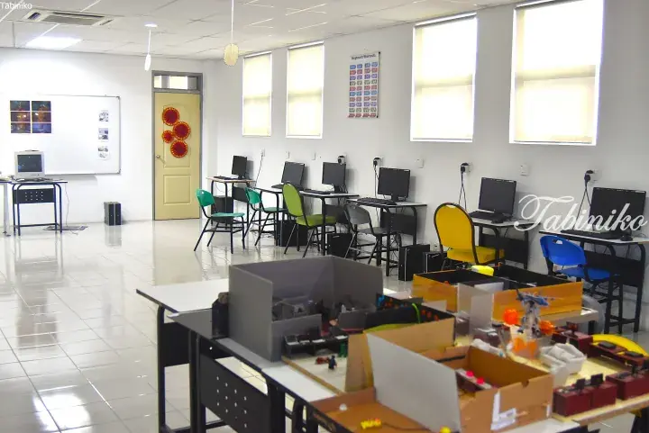 Bright computer lab with rows of PCs, colorful chairs, and a table displaying student robotics projects.