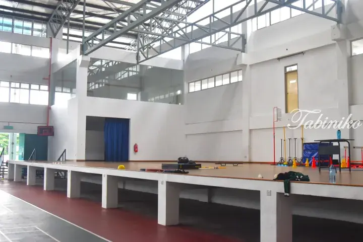 A spacious indoor gymnasium with a raised wooden stage, sports equipment, and high ceilings.