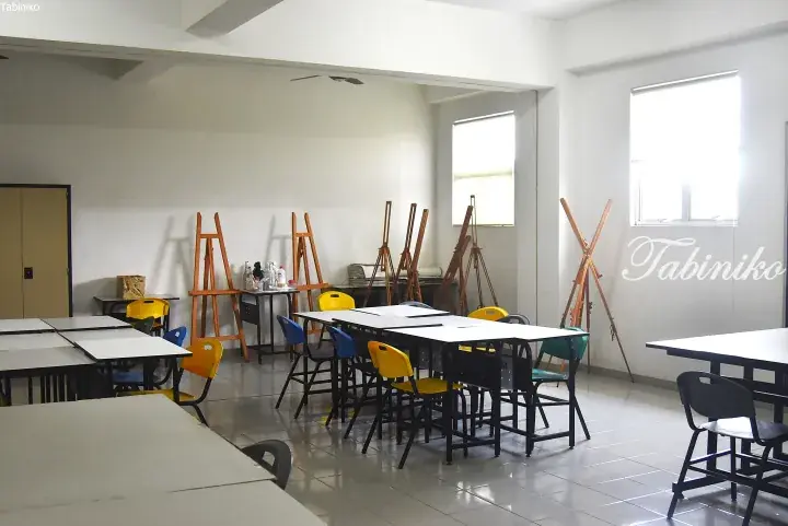 Bright art studio with wooden easels, white tables, and colorful chairs in a spacious room with large windows.