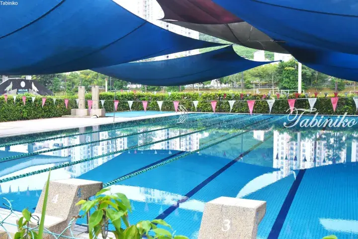 Swimming pool with lane ropes and starting blocks under blue shade sails at Nexus Wolf residential complex.