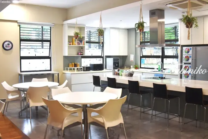 Modern shared kitchen with white tables, hanging plants, and stainless steel appliances in a Malaysian property.