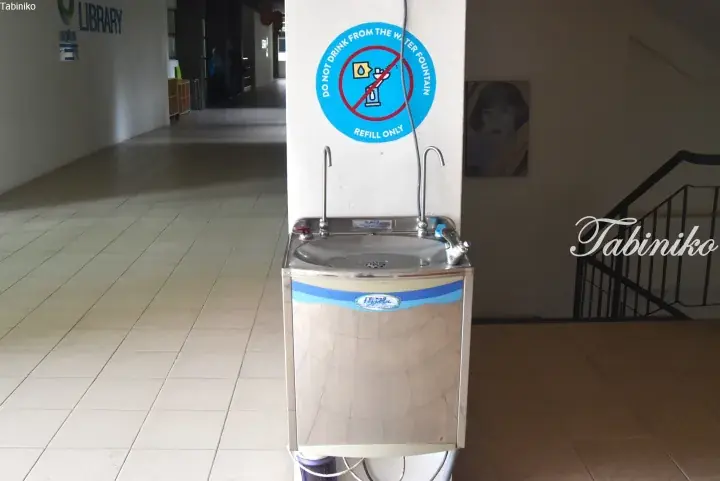Stainless steel water dispenser with a ‘Do Not Drink’ sign in a building hallway near a library.